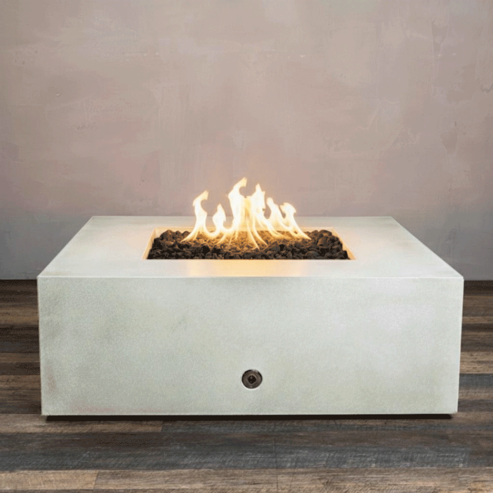 A studio close-up features a square fire pit in a crisp white finish, resting on a dark wood floor. Bright orange flames burn continuously from a bed of black lava rocks, contrasting with the smooth texture of the concrete fire pit. The visible chrome key valve on the side panel indicates the manual control of this natural gas fire pit unit, demonstrating its clean design and functionality.