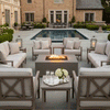 On a paved terrace beside a long rectangular pool in front of a brick manor, cushioned armchairs and sofas form a neat square around a pale brown square fire pit, its flame flickering over dark stones as lanterns, flower beds, and clipped hedges frame the scene, presenting a calm patio fire pit setup that works as a welcoming backyard fire pit focal point for relaxed evening drinks and soft outdoor entertaining near the water.