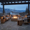 Beneath a dark pergola on a flagstone terrace overlooking snowy mountains, deep wood-framed sofas and chairs surround a broad brown concrete fire pit, its bright rectangular flame bed set into the center while lanterns, blankets, and steaming mugs rest on low tables, turning this elevated patio fire pit into a cozy backyard fire pit–style zone that naturally functions as a warm family gathering fire pit for conversation after time in the nearby hot tub.