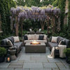 In a narrow garden court lush with ivy walls, potted greenery, and a pergola draped in purple wisteria, three gray cushioned sofas face a low brown square fire pit, its centered flames dancing above a bed of stones while lanterns and folded throws sit within reach, capturing a sheltered backyard fire pit nook that reads as a cozy backyard fire pit retreat designed for intimate outdoor entertaining and quiet evening chats.