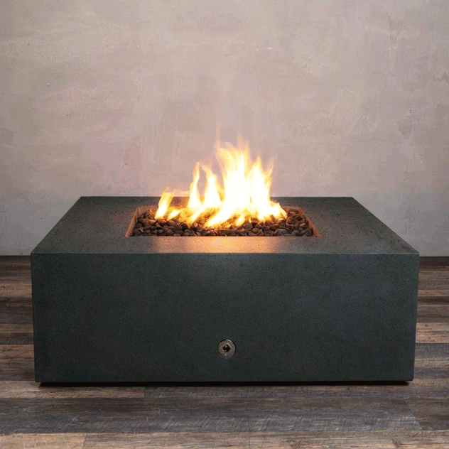 A deep graphite concrete fire pit with crisp square sides sits centered on a striped wood-look floor against a soft taupe wall, its recessed square opening filled with chunky brown lava rocks while bright yellow flames from the gas fire pit rise and sway in the middle, clearly revealing the broad top surface, small metal control on the front, and low profile of this compact square fire pit as it flickers steadily in place.