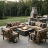 In a tree-lined backyard, cushioned wood armchairs and sofas form a loose square around a graphite square fire pit on a flagstone terrace, its lively flames dancing above dark rocks as lanterns, blankets, and a stone outdoor fireplace with grill station frame the scene, creating a patio fire pit seating area that feels like a warm backyard fire pit and natural family gathering fire pit for evening drinks and conversation beside the lawn.