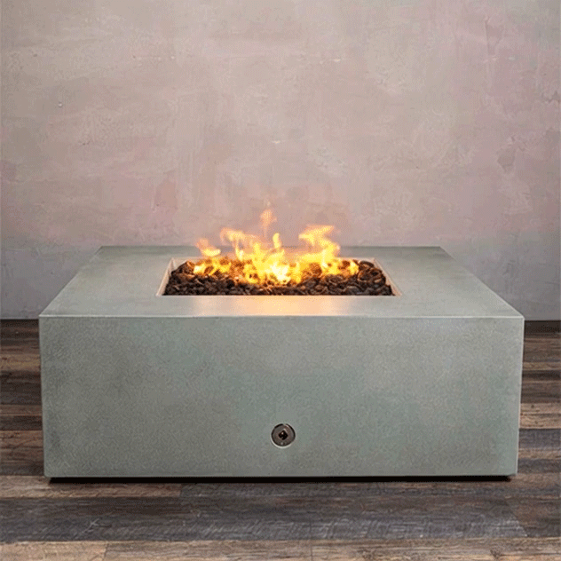A low, pale gray concrete fire pit with clean square edges sits centered on a striped wood-look floor against a soft taupe wall, its recessed square opening filled with dark lava rocks while bright yellow flames from the gas fire pit rise and fall in the middle, clearly showing the broad top surface, small base shadow, and compact proportions of this modern square fire pit as it flickers in place.