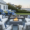 On a slate patio fire pit terrace beside a white coastal-style house, cushioned wood sofas with blue striped pillows frame a gray square fire pit whose flames dance above dark stones, while lanterns and drinks rest on a matching coffee table and lush hydrangeas border a lawn that leads to a harbor view with sailboats, capturing a relaxed backyard fire pit nook arranged for casual outdoor entertaining at sunset.