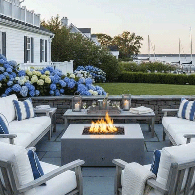 On a slate patio fire pit terrace beside a white coastal-style house, cushioned wood sofas with blue striped pillows frame a gray square fire pit whose flames dance above dark stones, while lanterns and drinks rest on a matching coffee table and lush hydrangeas border a lawn that leads to a harbor view with sailboats, capturing a relaxed backyard fire pit nook arranged for casual outdoor entertaining at sunset.