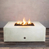 A pale stone-colored concrete fire pit with clean square sides sits centered on a striped wood-look floor against a soft taupe wall, its recessed square opening packed with dark lava rocks while tall yellow flames from the gas fire pit rise in the middle, clearly outlining the generous top surface, crisp edges, and compact proportions of this square fire pit as it flickers steadily in place.