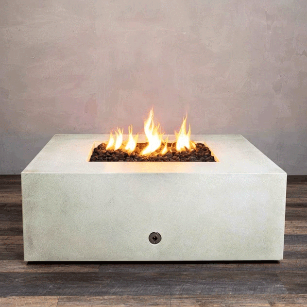 A pale stone-colored concrete fire pit with clean square sides sits centered on a striped wood-look floor against a soft taupe wall, its recessed square opening packed with dark lava rocks while tall yellow flames from the gas fire pit rise in the middle, clearly outlining the generous top surface, crisp edges, and compact proportions of this square fire pit as it flickers steadily in place.