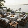 On a raised lakeside terrace, gray-cushioned wood armchairs form a circle around a low white square fire pit, its bright flame dancing above dark stones as lanterns, coffee cups, and a blanket-draped chair sit nearby, while a stainless grill, glass railing, and calm water with docks in the distance complete the scene, turning this patio fire pit area into a cozy backyard fire pit corner ideal for relaxed outdoor entertaining.