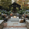 In a lush garden courtyard edged with stone walls and blooming hydrangeas, deep cushioned sectionals and chairs surround a white concrete fire pit with a square burner, its compact flame glowing over dark rocks as lanterns, pillows, and a basket of throws rest nearby, with a freestanding outdoor fireplace framed by trees behind, creating a sheltered backyard fire pit setting that naturally serves as a comfortable family gathering fire pit.