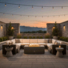 A large U-shaped sectional sofa wraps around a brown luxury fire pit on a stone patio overlooking mountain peaks at sunset. Overhead string lights enhance the ambiance of this backyard oasis, while stone walls provide privacy. The rectangular propane fire pit creates a warm central gathering point, perfect for a spacious family gathering fire pit arrangement in a scenic outdoor room.