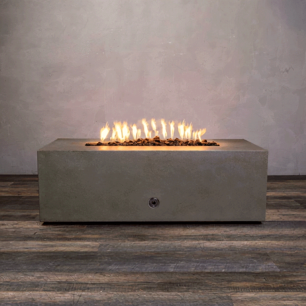 A studio view presents a rectangular concrete fire pit in a natural brown finish, resting on a dark wood floor against a textured beige wall. Bright orange flames rise continuously from a bed of black lava rocks along the length of the linear fire pit. The minimalist backdrop emphasizes the earth-tone texture and clean operation of a natural gas fire pit connection.