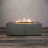 A low brown rectangular concrete fire pit burns with a continuous line of golden flames above small lava rocks, the modern fire pit centered on a wide plank floor against a smooth gray wall, and the GIF loops as the gas fire pit flickers gently, showing the clean edges, solid block shape, and narrow opening of this rectangular fire pit in a simple, uncluttered studio setting.