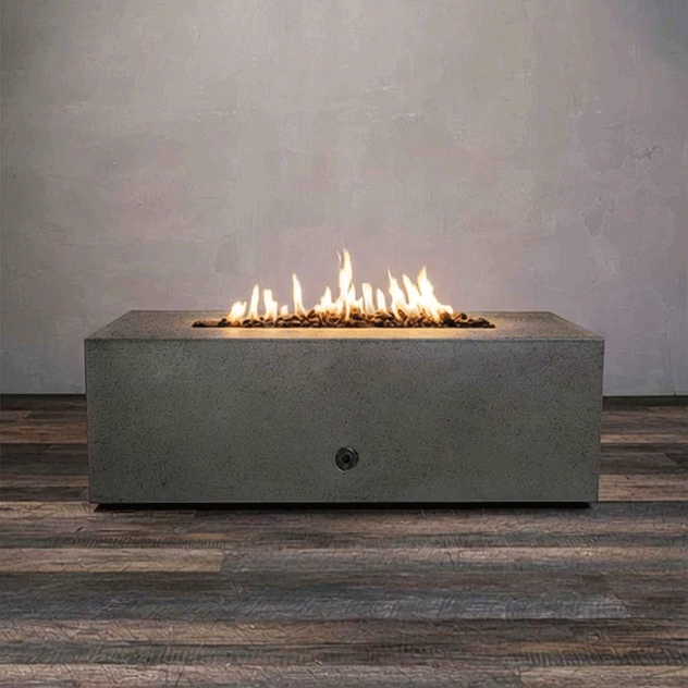 A low brown rectangular concrete fire pit burns with a continuous line of golden flames above small lava rocks, the modern fire pit centered on a wide plank floor against a smooth gray wall, and the GIF loops as the gas fire pit flickers gently, showing the clean edges, solid block shape, and narrow opening of this rectangular fire pit in a simple, uncluttered studio setting.