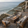 On a multi-level deck overlooking rolling ocean waves, a brown concrete fire pit flickers with a straight row of flames between modern wood-framed lounge chairs and a sofa, glass railings framing the view, planters and lanterns placed around, and wine glasses resting on nearby tables, the outdoor fire pit acting as a low fire table at the heart of this coastal seating area arranged for relaxed outdoor entertaining.