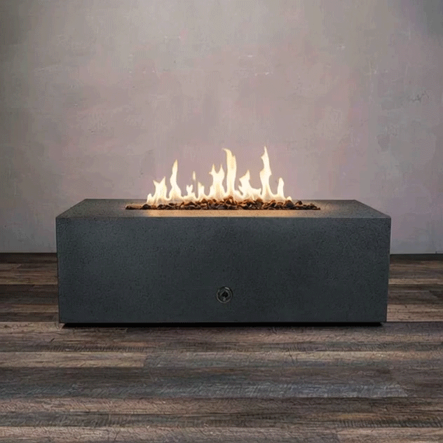 A dark graphite concrete fire pit with a long rectangular shape burns with a steady row of golden flames above small lava rocks, the modern fire pit centered on a wood plank floor against a plain gray wall, and the GIF loops as the gas fire pit quietly flickers, highlighting the clean edges, low profile, and narrow opening of this rectangular fire pit in a simple studio-style setting.