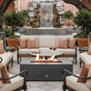 In a tiled courtyard framed by orange trees in large pots and a stone waterfall splashing into a pool, a graphite concrete fire pit glows with a line of flames between cream sofas with patterned coral pillows, the low patio fire pit set in front of a round mosaic table with wine glasses, acting as a sleek fire table that anchors this intimate seating area and enhances the scene as an inviting luxury fire pit lounge.