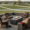 On a raised stone terrace overlooking rolling green pastures and white fences, a graphite concrete fire pit flickers in a narrow bed of lava rocks between leather armchairs and a tan sofa, riding boots and a helmet resting near rough stone side tables, the outdoor fire pit forming a central rectangle of flame that suggests a relaxed family gathering fire pit where people can unwind after time spent on the surrounding ranch.