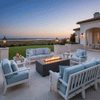 At sunset on a light stone patio beside a coastal-style home, a graphite concrete fire pit burns in a straight row of flames surrounded by white-framed sofas and chairs with soft blue cushions, lanterns glowing on the low walls, the patio fire pit centered in the seating group and helping define this open-air lounge as a refined backyard oasis that feels like a private luxury fire pit retreat facing the water.