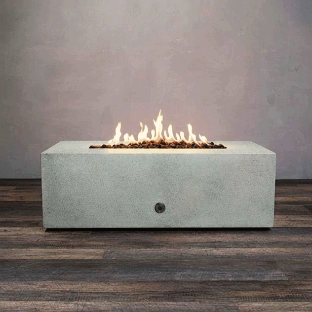 A low, light gray concrete fire pit with a clean rectangular shape burns in a narrow line of golden flames above smooth lava rocks, the modern fire pit centered on a wide plank floor against a plain gray wall, and the GIF loops as the gas fire pit quietly flickers, emphasizing the long opening, crisp edges, and minimalist profile of this rectangular fire pit in a simple studio setting.