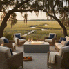 On an open patio overlooking marsh grasses and a winding creek at sunset, a rectangular concrete fire pit glows with a row of flames between woven lounge chairs with cream cushions and navy pillows, lanterns lined along the edge, and moss-draped branches arching overhead, the outdoor fire pit anchoring a relaxed seating circle that feels like a quiet backyard oasis designed for easy outdoor entertaining.