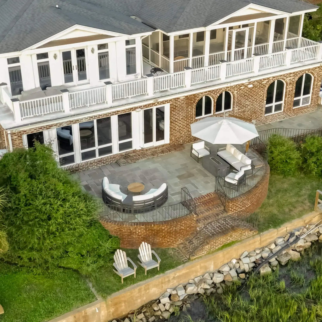 Aerial view of a stunning waterfront patio featuring a modern graphite-colored 55-inch rectangular Beton Concrete GFRC gas fire pit by Starfire Designs, surrounded by cozy white cushioned seating and a large cantilever umbrella, creating an inviting outdoor living space at a brick and white coastal home.