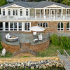 A stunning aerial view of a coastal backyard patio featuring the 55" Rectangle Beton Concrete GFRC Gas Fire Pit in Graphite by Starfire Designs. The sleek, modern fire pit sits center stage beneath a large white umbrella, surrounded by white-cushioned patio seating and set atop a curved brick terrace overlooking the waterfront. A serene setup perfect for relaxing by the water. Ideal for luxury outdoor living inspiration.