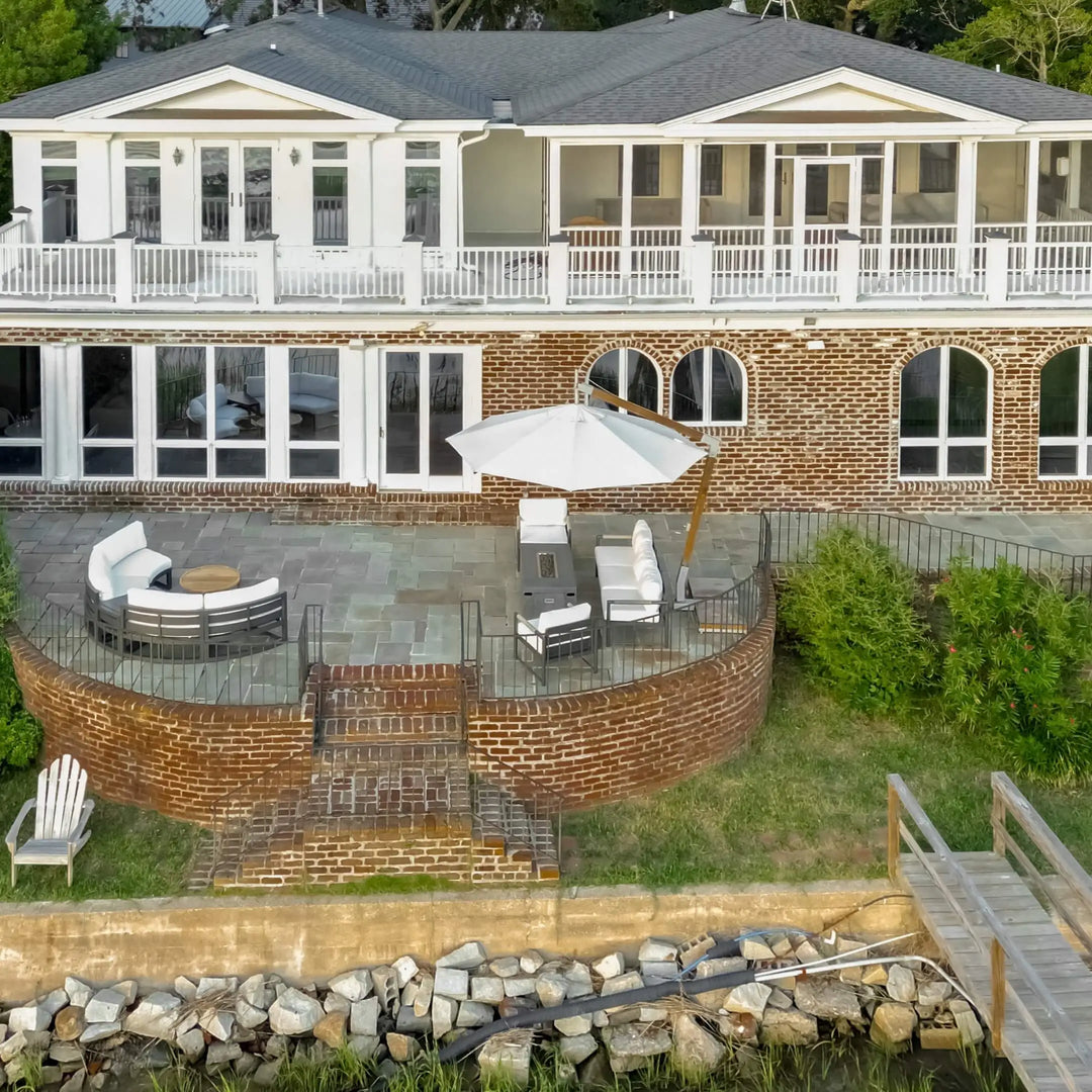 A stunning aerial view of a coastal backyard patio featuring the 55" Rectangle Beton Concrete GFRC Gas Fire Pit in Graphite by Starfire Designs. The sleek, modern fire pit sits center stage beneath a large white umbrella, surrounded by white-cushioned patio seating and set atop a curved brick terrace overlooking the waterfront. A serene setup perfect for relaxing by the water. Ideal for luxury outdoor living inspiration.