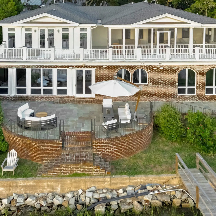 A stunning aerial view of a coastal backyard patio featuring the 55" Rectangle Beton Concrete GFRC Gas Fire Pit in Graphite by Starfire Designs. The sleek, modern fire pit sits center stage beneath a large white umbrella, surrounded by white-cushioned patio seating and set atop a curved brick terrace overlooking the waterfront. A serene setup perfect for relaxing by the water. Ideal for luxury outdoor living inspiration.