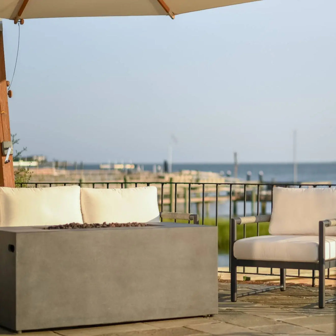 Modern patio scene featuring the Starfire Designs 55" Rectangle Beton Concrete GFRC Gas Fire Pit in Graphite finish, shown with the fire off. The clean, minimalist design pairs beautifully with cream-colored outdoor seating under a large umbrella, all set against a stunning coastal marina backdrop. Perfect for luxury backyard or waterfront entertaining.