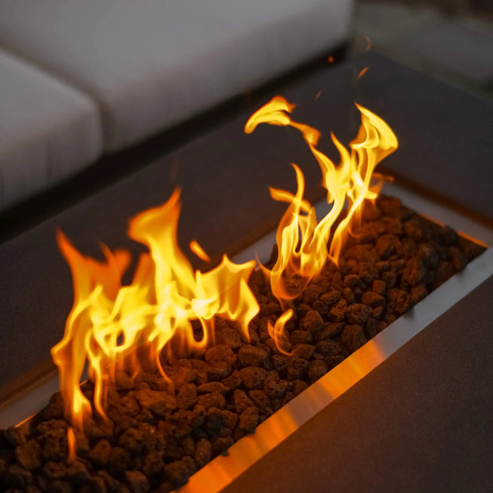 Close-up view of glowing flames dancing over lava rocks in the 55-inch Rectangle Beton Concrete GFRC Gas Fire Pit by Starfire Designs, featuring a hidden propane tank and graphite finish, set on a cozy patio with a lakefront backdrop / perfect for outdoor gatherings and modern relaxation.