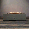 A studio presentation displays a rectangular fire pit in a natural brown finish, resting on a dark wood floor. Bright yellow flames burn continuously along the linear fire pit burner, contrasting with the earth-tone texture of the concrete fire pit. The visible chrome key valve on the side panel suggests a manual ignition system, demonstrating the clean design of this natural gas fire pit unit.