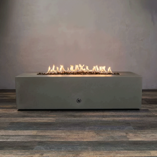 A studio presentation displays a rectangular fire pit in a natural brown finish, resting on a dark wood floor. Bright yellow flames burn continuously along the linear fire pit burner, contrasting with the earth-tone texture of the concrete fire pit. The visible chrome key valve on the side panel suggests a manual ignition system, demonstrating the clean design of this natural gas fire pit unit.