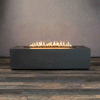 A studio presentation highlights a rectangular fire pit in a matte graphite finish, resting on a dark wood floor. Bright yellow flames burn continuously along the linear fire pit burner, contrasting with the smooth texture of the concrete fire pit. The visible chrome key valve on the side panel indicates the manual control of this natural gas fire pit unit, demonstrating its clean design and functionality.