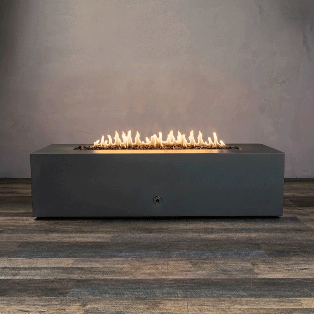 A studio presentation highlights a rectangular fire pit in a matte graphite finish, resting on a dark wood floor. Bright yellow flames burn continuously along the linear fire pit burner, contrasting with the smooth texture of the concrete fire pit. The visible chrome key valve on the side panel indicates the manual control of this natural gas fire pit unit, demonstrating its clean design and functionality.