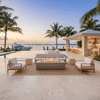 A tropical poolside cabana features a gray luxury fire pit positioned on a travertine patio. Surrounded by white cushioned sofas and armchairs, the unit overlooks an infinity pool and docked yacht at sunset. The warm glow of the fire table creates an inviting atmosphere for outdoor entertaining, anchoring the spacious patio fire pit arrangement.