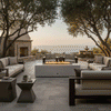 A stone terrace overlooking the ocean features a white luxury fire pit surrounded by sofas and armchairs. Large oak trees and an outdoor fireplace frame the scene, creating an inviting family gathering fire pit area. The warm flames of the fire feature serve as a central anchor for the outdoor fire pit arrangement at sunset.