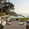 A white modern fire pit sits on a concrete patio next to an infinity pool, overlooking a hillside and ocean view. Surrounded by contemporary furniture, the patio fire pit anchors the outdoor lounge area. The clean-burning flames of the smokeless fire pit enhance the serene atmosphere, utilizing a gas fire pit connection for a seamless look.