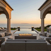 A grand estate loggia features a white fire table positioned between large sofas, overlooking an infinity pool and ocean horizon. The setting is framed by stone arches and columns, creating a luxurious backyard oasis. The steady flames of the propane fire pit provide warmth and ambiance for outdoor entertaining in a sophisticated covered space.