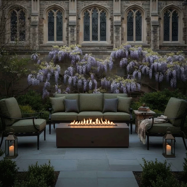 In a walled courtyard framed by old stone architecture and cascading purple wisteria, a long rectangular concrete fire pit blazes in a narrow channel of flames between deep green cushioned patio seating, the patio fire pit resting on smooth gray pavers with candle lanterns set nearby, and the GIF lingers on the warm glow of this linear fire feature as the concrete fire pit anchors the space and invites quiet conversation in the tranquil outdoor setting.
