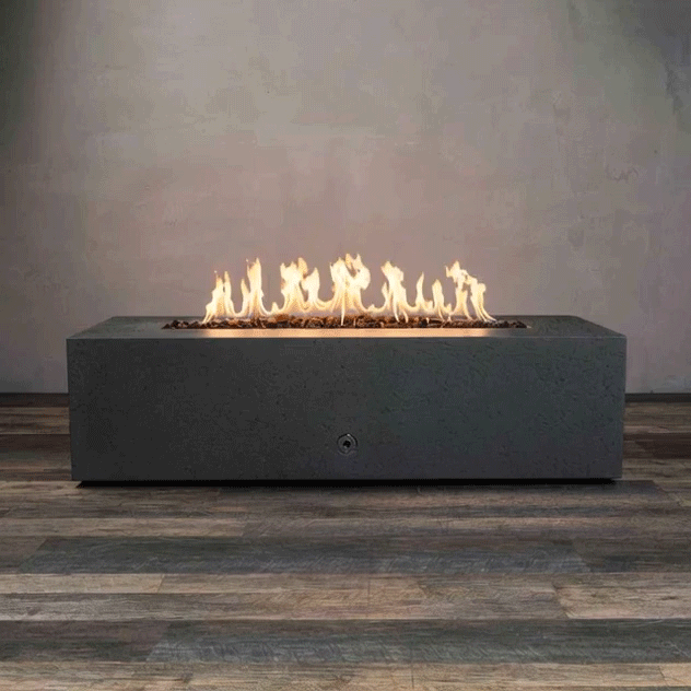 A graphite concrete fire pit with clean rectangular sides sits centered on a wood-look floor in a bare room, its recessed trough filled with dark lava rocks while a bright ribbon of yellow flames dances along the burner, emphasizing the low, boxy silhouette of this concrete fire pit and the long, horizontal line of a sleek rectangular fire pit that reads as a minimalist linear fire pit and modern fire pit design.