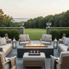 On a broad stone terrace overlooking an expansive lawn and distant water, a graphite concrete fire pit stretches between woven armchairs and sofas, its long burner sending up a continuous row of flames above dark stones while tall lanterns and clipped shrubs flank the seating, presenting a substantial concrete fire pit that feels like a luxury fire pit and refined family gathering fire pit at the heart of an elegant backyard oasis.