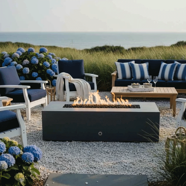 A graphite concrete fire pit glows on a gravel patio nestled among tall beach grasses and bright blue hydrangeas, its linear flames rising above lava rocks as white and navy coastal-style chairs and a matching sofa circle around, wine glasses and trays on the low table nearby, creating an outdoor fire pit scene where the sturdy concrete fire pit becomes a cozy backyard fire pit in a breezy seaside backyard oasis.