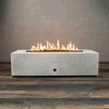 A long white concrete fire pit with crisp rectangular sides sits centered on a wood-look floor against a plain wall, its recessed channel filled with dark lava rocks while a bright ribbon of flames runs the full length of the burner, highlighting the low, boxy shape of this concrete fire pit and the clean lines of a modern rectangular fire pit with a gently flickering linear fire pit flame pattern.