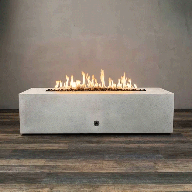 A long white concrete fire pit with crisp rectangular sides sits centered on a wood-look floor against a plain wall, its recessed channel filled with dark lava rocks while a bright ribbon of flames runs the full length of the burner, highlighting the low, boxy shape of this concrete fire pit and the clean lines of a modern rectangular fire pit with a gently flickering linear fire pit flame pattern.