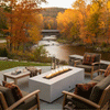 On a stone patio beside a gently curving river framed by vivid autumn trees, a white concrete fire pit burns with a long, even line of flame between cushioned wooden chairs, plaid pillows, and a low table holding mugs and lanterns, creating an outdoor fire pit scene where the sturdy rectangular form becomes a cozy backyard fire pit and inviting family gathering fire pit for crisp fall evenings.