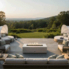 A broad terrace overlooking rolling hills and distant forest features a white concrete fire pit at its center, the rectangular opening packed with dark stones and a continuous flame while woven sofas, round daybeds, and soft pillows curve around it, turning this concrete fire pit into an elegant outdoor fire pit and rectangular fire pit focal point that anchors a serene backyard oasis for lounging at sunset.
