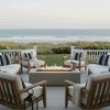 On a raised coastal deck with white railings and views of dunes and ocean waves, a low white concrete fire pit glows between wood-framed chairs and sofas upholstered in cream cushions with navy striped pillows, blankets and a champagne bucket nearby, showing a patio fire pit that functions as a sleek concrete fire pit centerpiece for relaxed outdoor entertaining in a breezy beachside backyard oasis.
