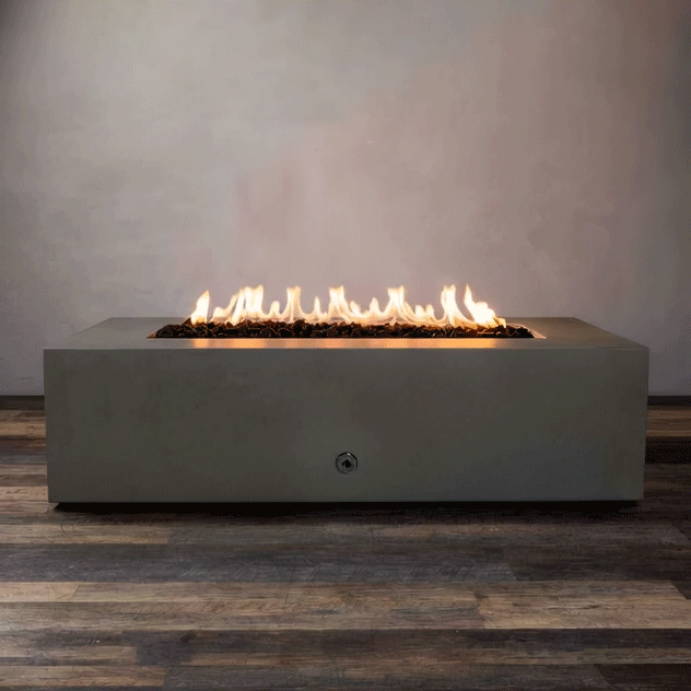 A taupe concrete fire pit with crisp, low walls sits centered on a mixed-tone wood floor, its long recessed trough filled with dark lava rocks while a steady ribbon of flame from the gas fire pit rises and flickers across the length of the rectangular fire pit, showing a clean control port on the front panel and emphasizing the simple blocky shape of this modern fire pit against a softly textured light gray wall.