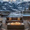 On a wide stone terrace high in the snowy mountains, a sleek taupe concrete fire pit burns in a long narrow channel between deep gray sofas and chairs, its lively flames standing out against nearby snow patches, a steaming spa, and dramatic peaks in the distance, turning the modern fire pit into a welcoming outdoor fire pit focal point that naturally works as a family gathering fire pit for après-ski relaxation and stargazing.