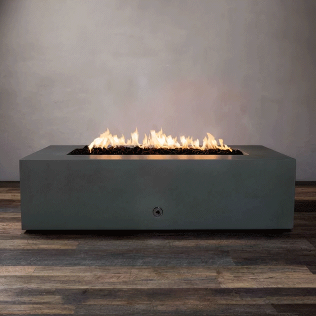 studio presentation displays a rectangular fire pit in a matte graphite finish, resting on a dark wood floor. Bright yellow flames burn continuously along the linear fire pit burner, contrasting with the smooth texture of the concrete fire pit. The visible chrome key valve on the side panel suggests a manual ignition system, demonstrating the clean design of this natural gas fire pit unit.